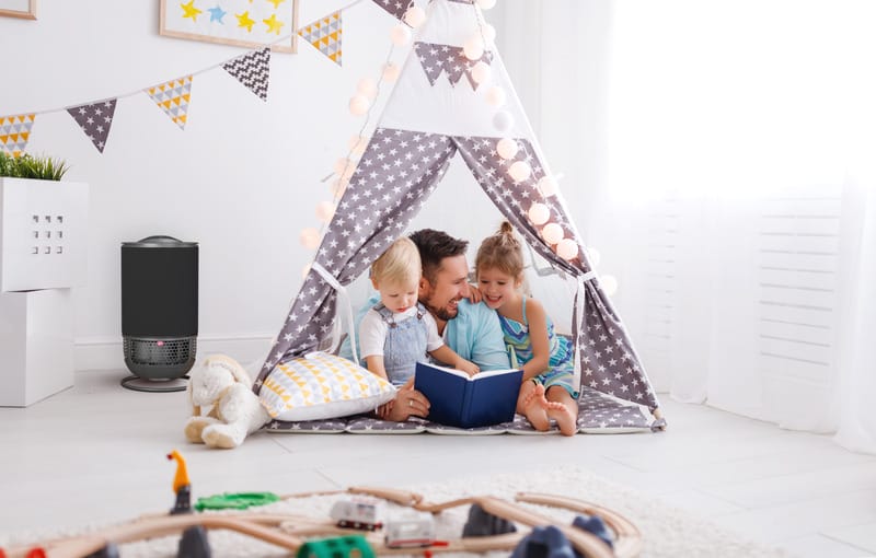 Aeroguard 360 PRO & Premium A father sits inside a star-patterned play tent with two young children, reading a book together. The cosy, well-lit room features playful decor, wooden train tracks, and an Aeroguard 360 PRO & Premium air purifier by Lux for fresh air.