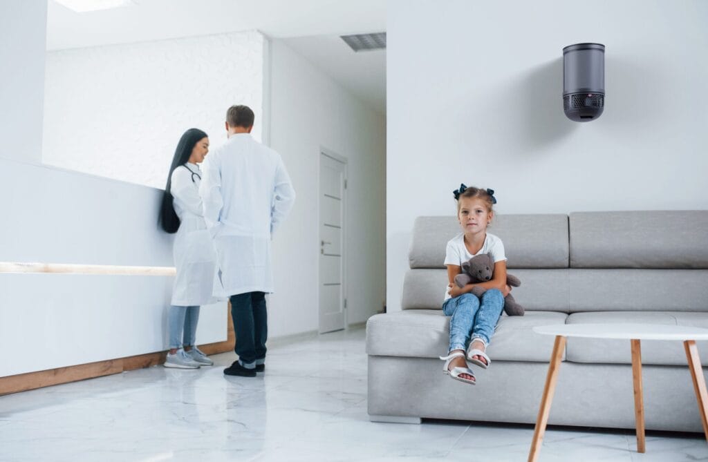Aeroguard 360 PRO & Premium A young girl holding a teddy bear sits on a grey sofa in a modern, white-walled waiting room. Two adults in lab coats talk near a counter, while an air purifier by Lux, Aeroguard 360 PRO & Premium ensures the air stays fresh and clean.