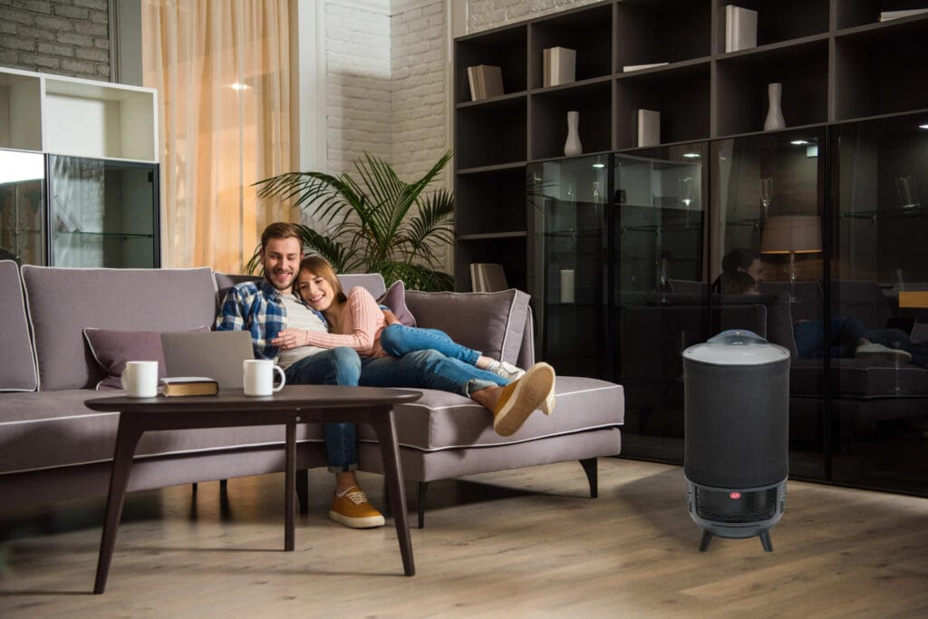 Aeroguard 360 PRO & Premium A smiling couple sits on a grey sofa in a modern living room, cuddling and looking at a laptop. Nearby on the wooden floor is an Aeroguard 360 PRO & Premium air purifier by Lux, adding comfort to their cosy space.