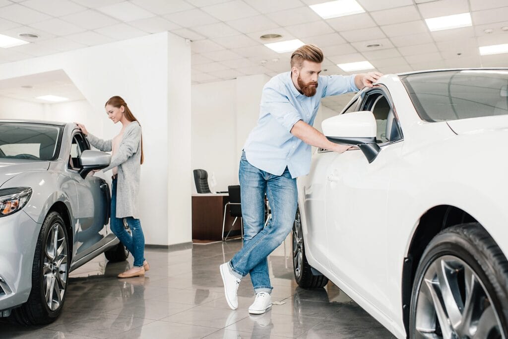 Aeroguard 360 PRO & Premium A man inspects a white car while leaning on its wing mirror in a showroom, and a woman looks inside a silver car in the background. Both appear to be considering vehicles for purchase.