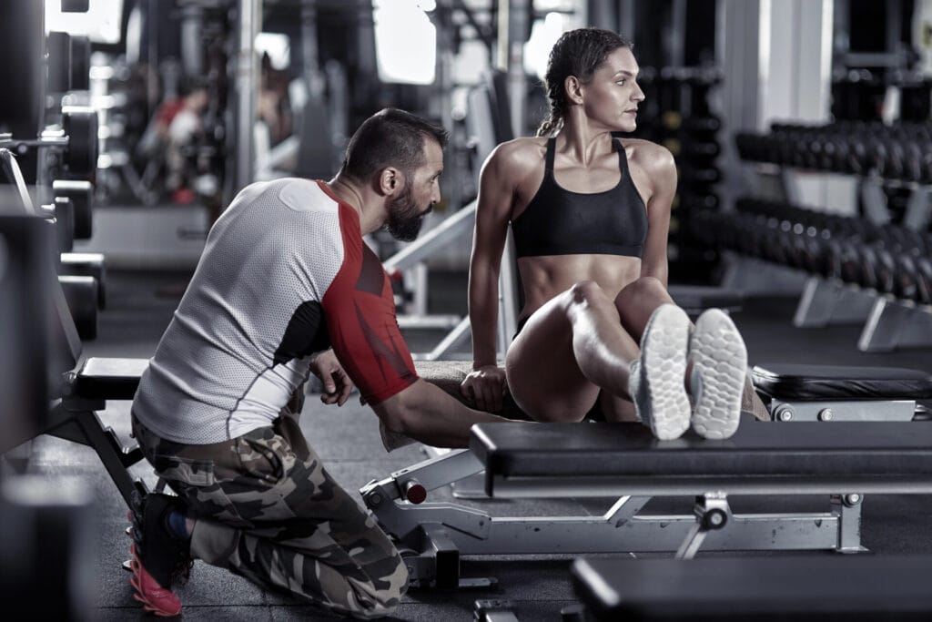 Aeroguard 360 PRO & Premium A man helps a woman in sportswear perform a tricep dip exercise on a bench in a gym filled with weights and fitness equipment.