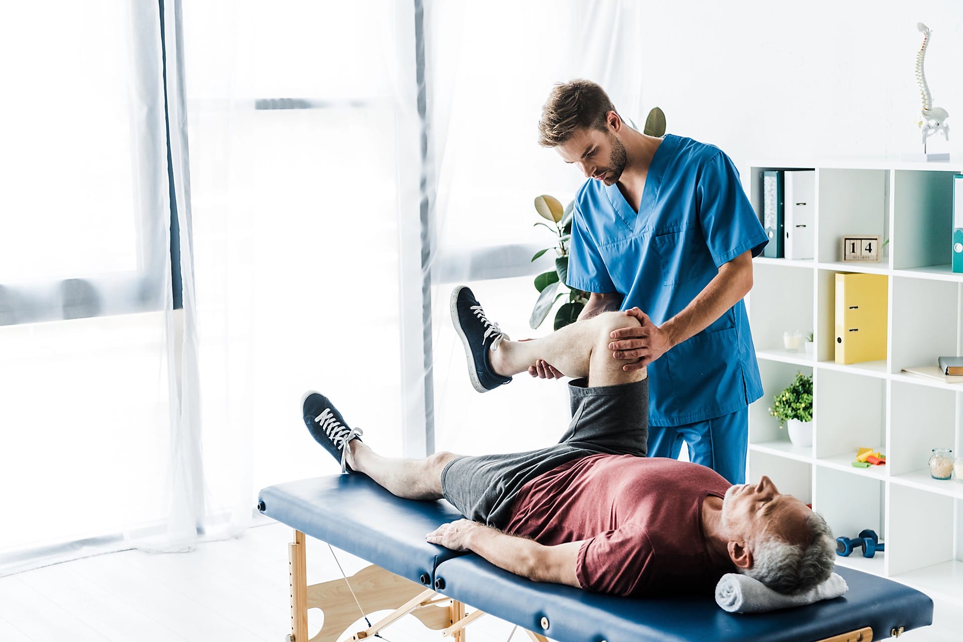 Aeroguard 360 PRO & Premium A physiotherapist in blue scrubs helps an older man lying on a treatment table by stretching his leg in a bright, modern clinic. Shelves and a window are visible in the background.