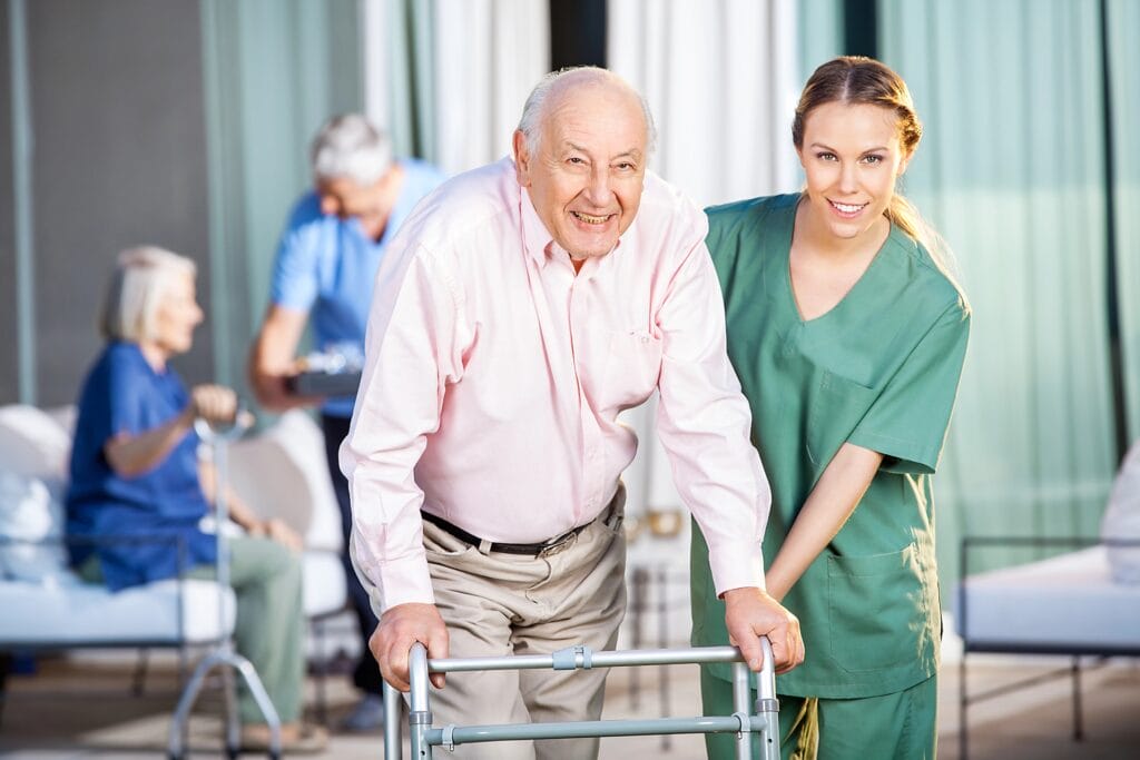 Aeroguard 360 PRO & Premium An elderly man using a zimmer frame smiles beside a carer in green scrubs. In the background, another older woman sits with a zimmer frame, and a man stands nearby. The setting appears to be a care home.