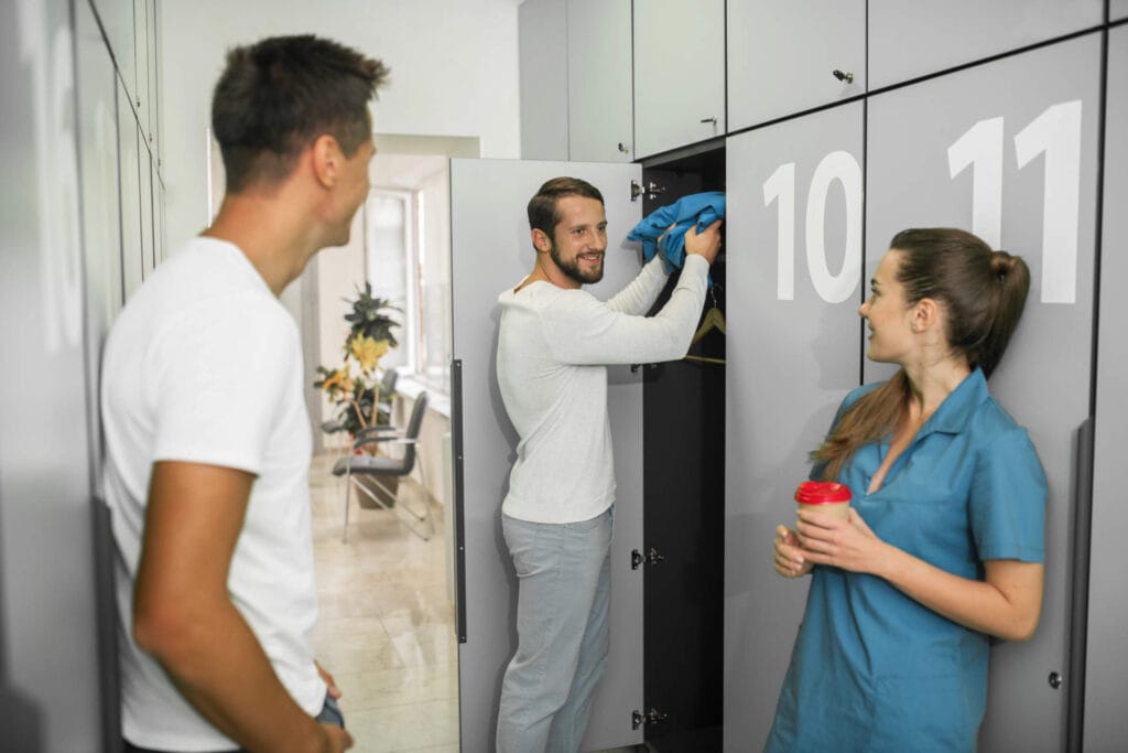 Aeroguard 360 PRO & Premium Three people stand near lockers in a corridor. One man is putting clothes into a locker, another man in a white shirt is talking to him, and a woman in blue scrubs holds a red cup, smiling.